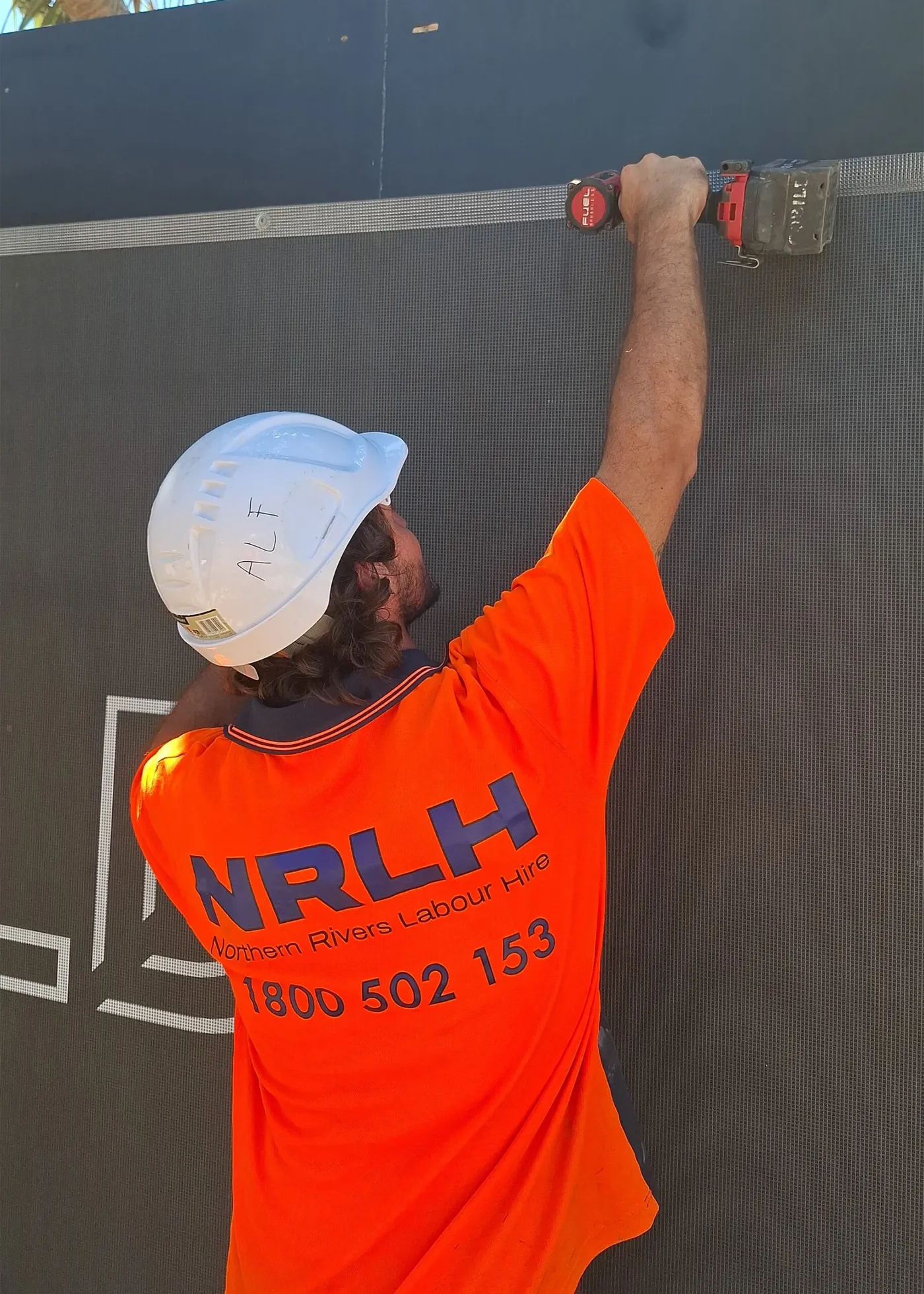 An NRLH labourer in a white hard hat fixing site hoarding overhead