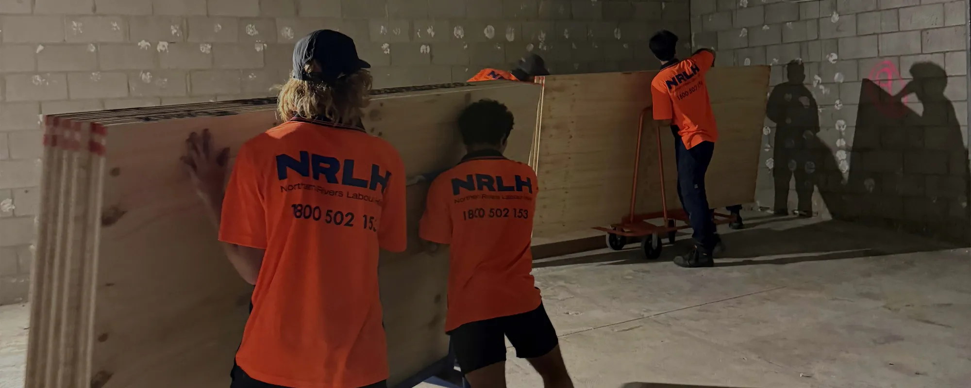 Three NRLH labourers carrying sheets of ply inside a blockwork room